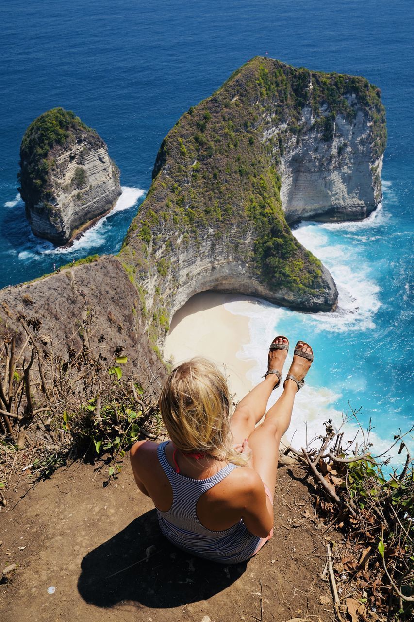 REAR VIEW OF WOMAN SITTING ON ROCK AT SHORE