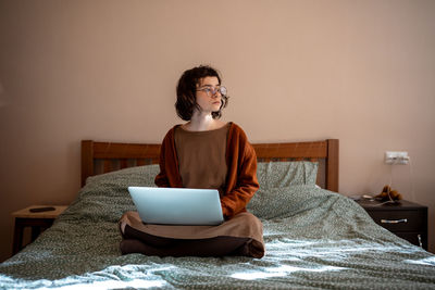 Teen girl student studying on laptop sitting in bed at home looking at window.