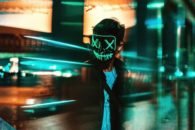 Man wearing neon mask with reflection of illuminated city on glass window at night