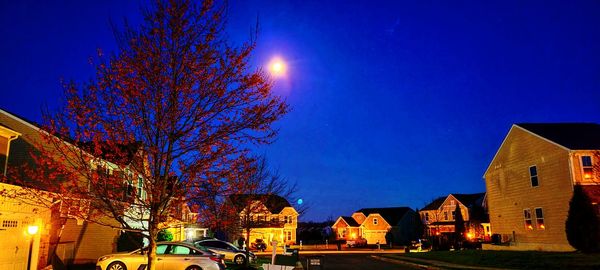 Illuminated street by buildings against blue sky at night