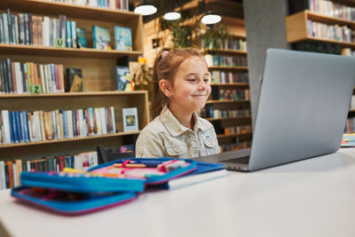 Pleased little student having video class remotely with her teacher on laptop sitting in library