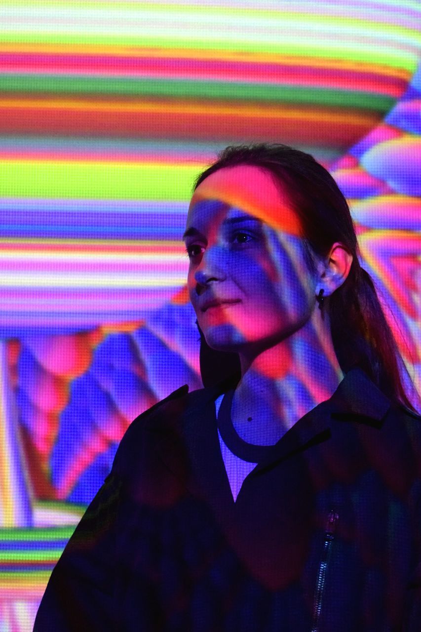 PORTRAIT OF A YOUNG WOMAN LOOKING AWAY AT ILLUMINATED NIGHTCLUB