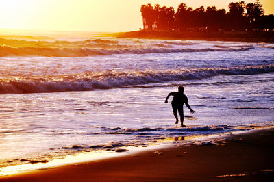 Silhouette man running with surfboard on beach against sky during sunset