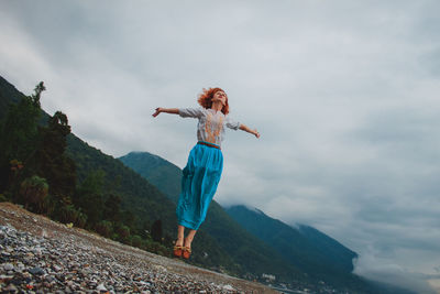 Low angle view of woman levitating over field by mountain against cloudy sky
