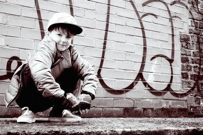 Low angle portrait of smiling boy crouching on sidewalk against wall