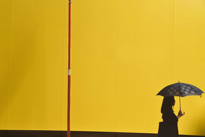 Side view of mature woman with umbrella walking by yellow wall