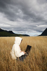 Mid adult woman using laptop while sitting on grassy field against cloudy sky