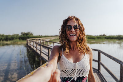 Woman holding hand of boyfriend while standing on footbridge against sky