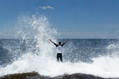 Rear view of man standing by waves splashing at shore against sky