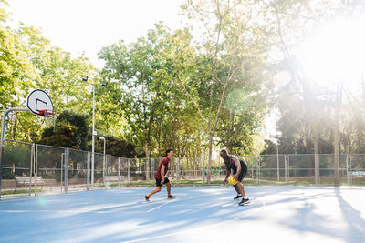 Side view of full body multiracial friends players running together with ball while playing basketball on sports ground in sunny day
