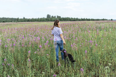 Rear view of young blond woman walking on field among flowers fireweed with dog, traveling with pets