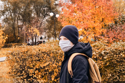 Boy wearing mask looking away while standing against autumn plants