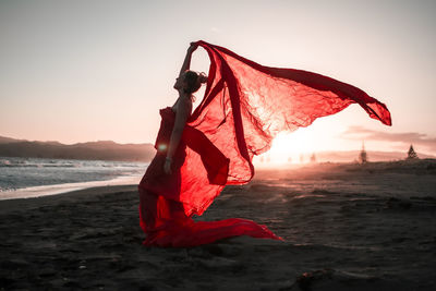 Young woman wearing red dress standing on beach against sky during sunset