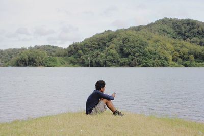 Rear view of man sitting by lake against trees