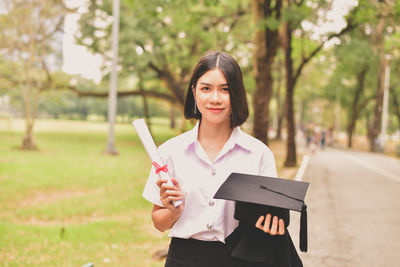 Portrait of young woman holding mortarboard while standing at park
