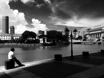 Full length of man sitting by river against sky in city