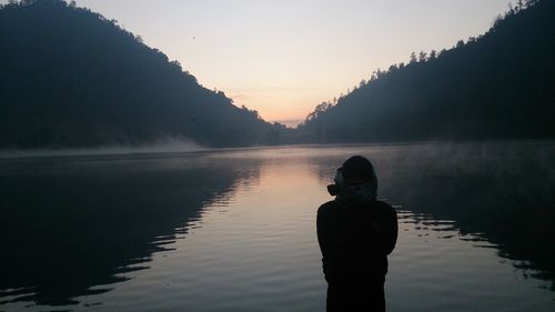 Rear view of man standing by lake against sky during sunset