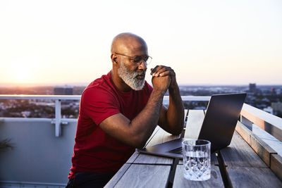 Mature bearded man looking at laptop while sitting on building terrace in city during sunset