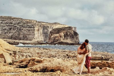 Rear view of man and a woman standing on rock by sea against sky