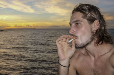 Close-up of shirtless man holding cigarette against sky during sunset