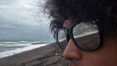 Close-up of young man wearing sunglasses at beach against cloudy sky