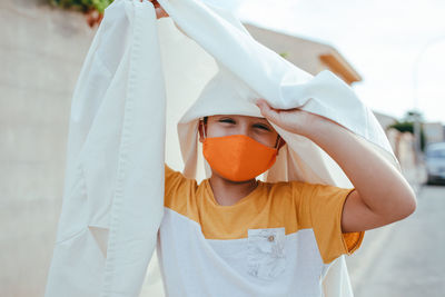 Delighted boy in ghost costume and protective mask from coronavirus standing in city during halloween holiday and looking at camera