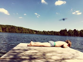 Full length of shirtless boy resting on pier over lake against sky