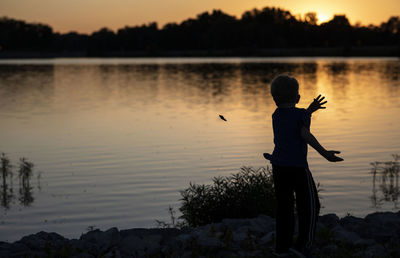 Rear view of silhouette man standing at lakeshore during sunset
