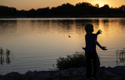 Rear view of silhouette boy throwing stone in lake during sunset