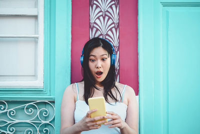 Shocked woman using mobile phone while listening music against wall