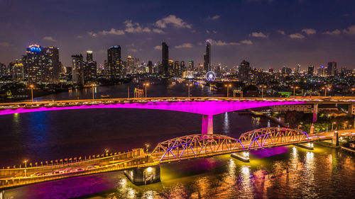 Illuminated bridge over river by buildings against sky at night