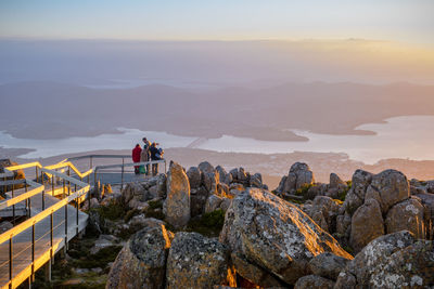 People on rocks by mountains against sky during sunset