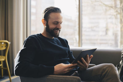 Happy businessman using digital tablet while sitting on sofa in office lobby