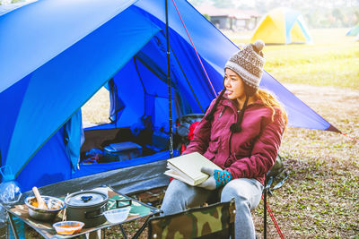 Young woman reading book while sitting on chair at campsite