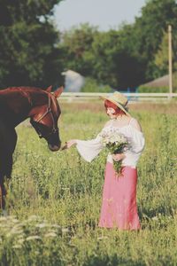 Full length of woman wearing hat feeding horse on field