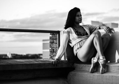 Side view of thoughtful young woman sitting on building terrace against sky