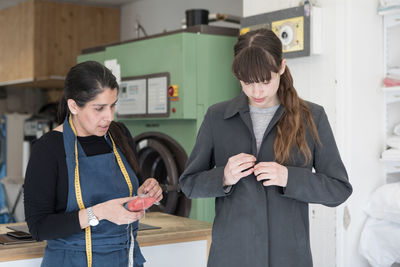 Mature female tailor standing by young customer wearing overcoat at laundromat