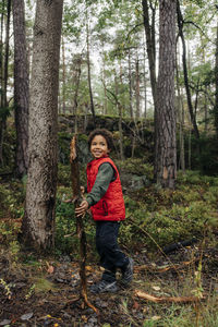 Side view of boy looking away holding firewood by trees in forest