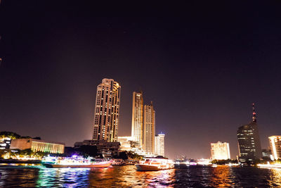 Illuminated modern buildings by river against sky at night