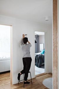 Full length of woman hammering nail on wall while standing on stool at home