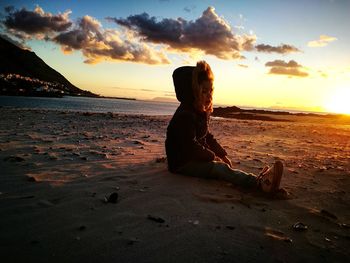 Man sitting on shore at beach against sky during sunset