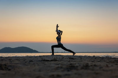 Side view of woman with arms raised exercising at beach against sky during sunset