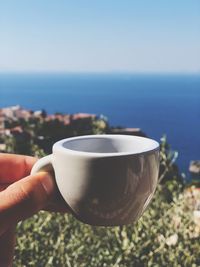 Cropped image of hand holding coffee cup against sea and sky
