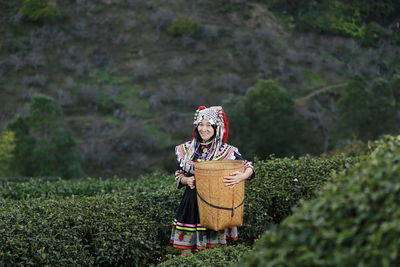 Portrait of smiling woman with wicker basket standing amidst tea crops on farm