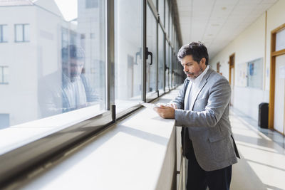 Professor using digital tablet standing by window sill in corridor at university