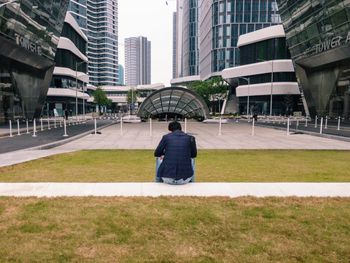 Rear view of man sitting against modern buildings in city