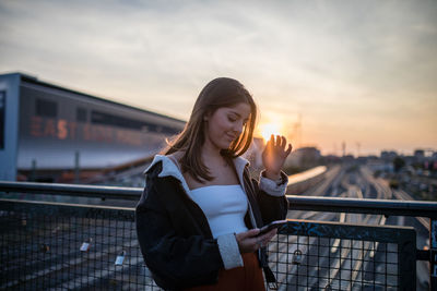 Woman using mobile phone while standing on bridge against railroad tracks during sunset