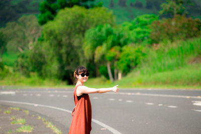 Side view of young woman wearing sunglasses hitchhiking while standing on road