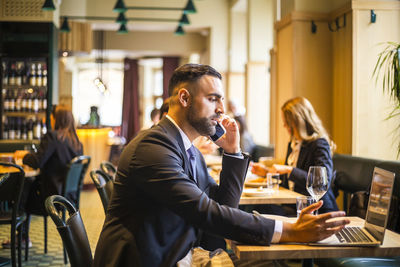 Side view of businessman talking on phone while using laptop in restaurant