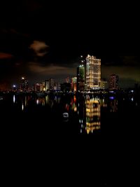 Illuminated modern buildings by sea against sky at night